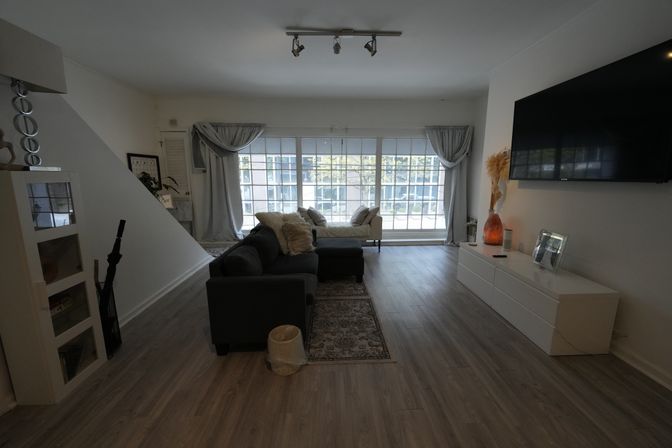 Modern living room with gray sectional and chaise, large multi-pane windows with curtains, light wood floors, area rug, wall-mounted TV and white media console.