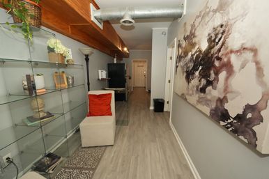 Modern apartment hallway interior with glass display shelves, a beige ottoman with a red pillow, exposed metal ductwork beneath a wood beam, light wood floors, and oversized abstract wall art.
