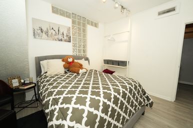 Cozy modern apartment bedroom with gray quatrefoil duvet, plush teddy bear on pillows, upholstered headboard, city skyline wall art, glass block accent and open shelving with storage bins.