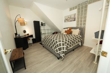 Cozy small apartment bedroom with gray upholstered bed, patterned duvet and teddy bear, desk and lamp, glass shelving and wood‑look laminate flooring under a sloped ceiling.