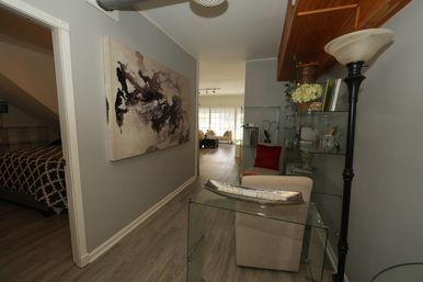 Bright modern hallway with gray walls and wood-look flooring, abstract canvas art, glass shelves and console, a beige armchair with red pillow, leading to a sunlit living room with large windows.