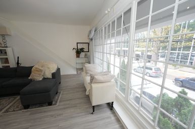 Sunlit street-facing living room with large grid windows, gray L-shaped sofa with plush throw, cream armchair with decorative pillows, light gray wood floors and view of parked cars and trees outside.