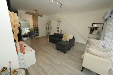 Bright, airy living room with a dark gray sectional and chaise on pale wood floors, glass-block accent wall, white walls, large windows with sheer curtains, potted plants and neutral pillows.