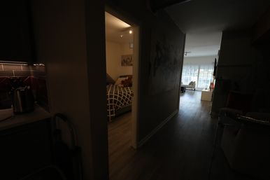 Dim apartment hallway with wood floors leading past a cozy bedroom with patterned bedspread and a teddy bear to a bright living room with large windows; kitchen counter and kettle visible at left.