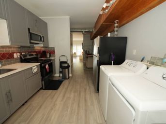 Compact galley kitchen with light wood laminate floor, gray cabinets, red-and-gray tile backsplash, stainless steel stove and refrigerator, trash can, and side-by-side white washer and dryer.