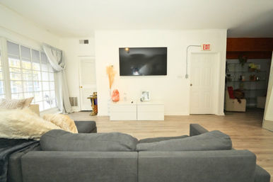 Sunlit modern living room with a gray sectional sofa facing a wall-mounted flat-screen TV above a white media console, large floor-to-ceiling windows with light curtains, pale wood floors and an exit door on the right.