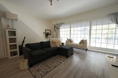 Sunlit modern living room with floor-to-ceiling grid windows, dark gray sectional sofa and ottoman, cream daybed, light wood floors and decorative rug.