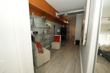 Modern urban loft hallway with glass display shelves, beige ottomans with red pillows, exposed metal ductwork and wood beam over gray laminate floors leading to a compact kitchen and living area.