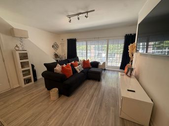 Cozy modern apartment living room with large grid window, black sectional sofa with orange pillows, light wood laminate floors, white media console and wall-mounted TV.