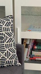 Cozy living-room corner with a navy-and-white geometric throw pillow on a gray sofa beside a white glass-front cabinet filled with stacked books, including a visible Spanish dictionary.