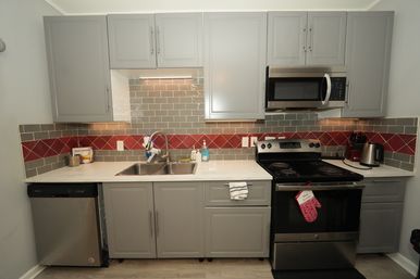 Bright modern gray kitchen with stainless steel oven, microwave and dishwasher, double-basin sink under gray cabinets, white countertop and gray subway tile backsplash with a red diagonal accent stripe, electric cooktop, kettle and knife block.
