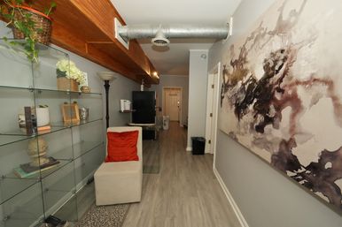 Cozy modern apartment hallway with glass shelving, potted plants, a beige chair with bright orange cushion, large abstract wall art, exposed metal ductwork and wood ceiling beam over gray laminate floors.
