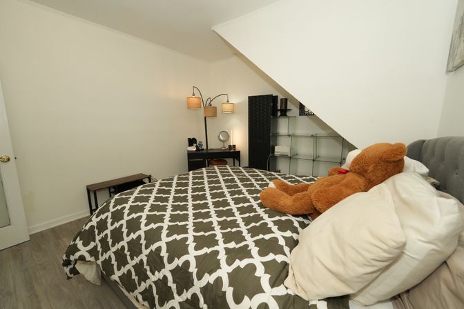 Cozy sloped-ceiling bedroom with gray-and-white geometric duvet, large plush teddy bear on pillows, tufted headboard, corner desk with arc lamp and glass shelving under the stairs
