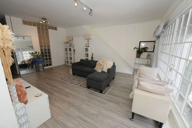 Bright modern living room with gray sectional, light wood laminate floors, large wall of windows, glass-block accent wall, plants and cozy neutral decor.