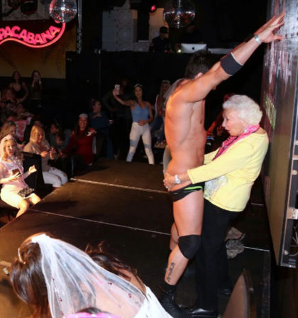 Male stripper on a nightclub stage with an older woman playfully grabbing him during a bachelorette party, cheering women and disco ball in the background