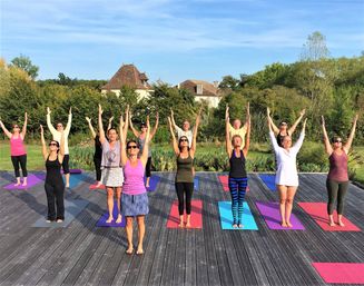 Sunlit outdoor yoga class on a wooden deck in a rural estate setting, adults on colorful mats standing with arms raised toward a blue sky, surrounded by trees and a stone house in the background.
