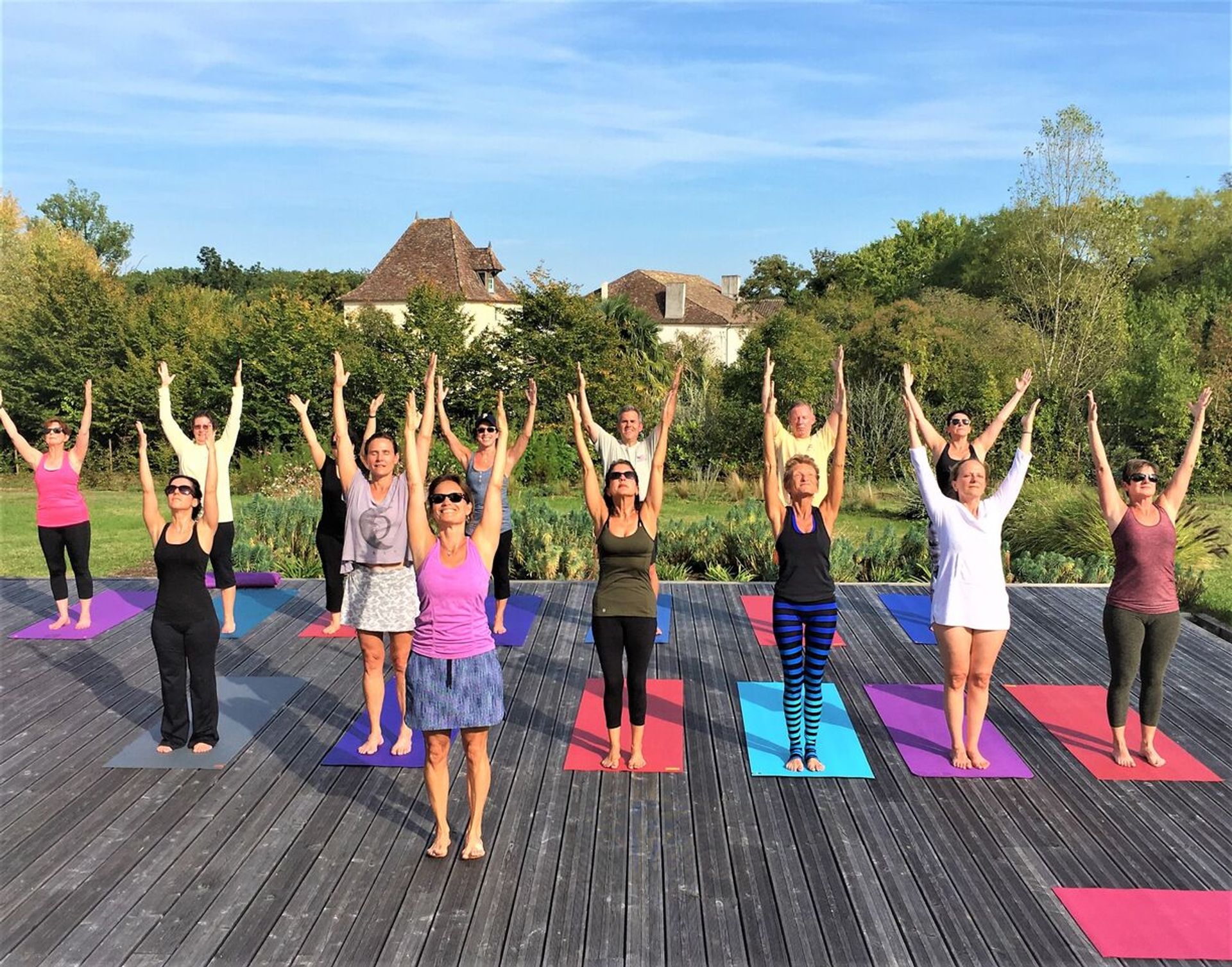 Sunlit outdoor yoga class on a wooden deck in a rural estate setting, adults on colorful mats standing with arms raised toward a blue sky, surrounded by trees and a stone house in the background.