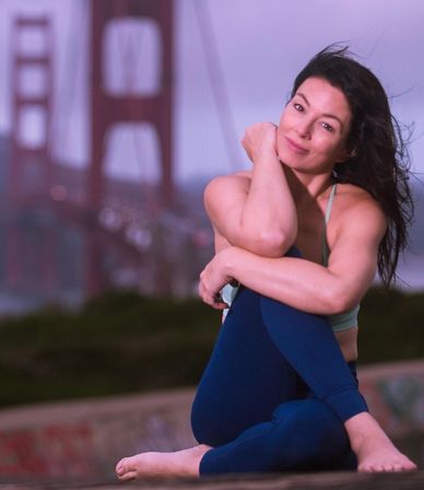 Smiling woman in sports bra and leggings seated in a relaxed yoga pose, windblown hair, Golden Gate Bridge and San Francisco skyline blurred behind her at dusk