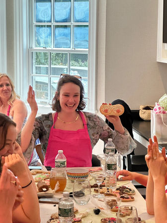Smiling woman in a pink apron holds a decorated cookie at a sunlit indoor cookie-decorating party, surrounded by friends, drinks, and baking supplies.
