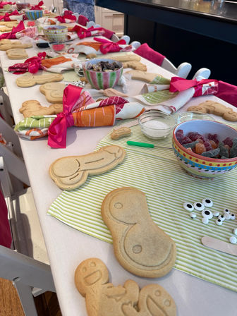 Long home kitchen table set up as a kid-friendly DIY cookie-decorating station with un-iced sugar cookies, bowls of gummy candy and candy eyes, striped placemats and colorful napkins tied with pink ribbons.