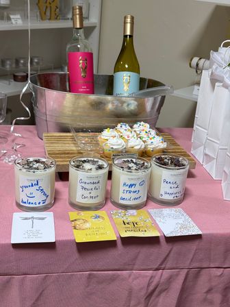 Indoor celebration table with metal ice tub holding two colorful wine bottles, a tray of frosted sprinkle cupcakes, four handmade scented candles with handwritten affirmation labels and matching inspirational cards, arranged on a pink tablecloth next to white gift bags.