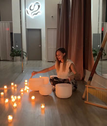Person seated in a calm wellness studio playing white crystal singing bowls for a sound bath meditation, surrounded by glowing votive candles and warm studio lighting.