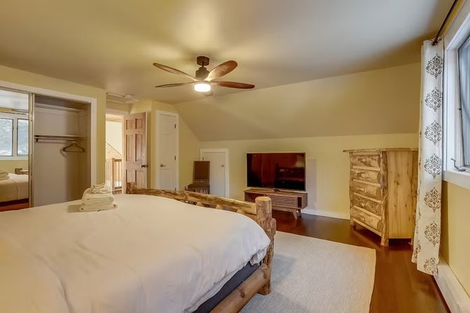 Cozy rustic bedroom with log-frame bed topped with folded towels, white bedding, ceiling fan, flat-screen TV on a media console, wooden dresser, hardwood floors and patterned curtain by a window