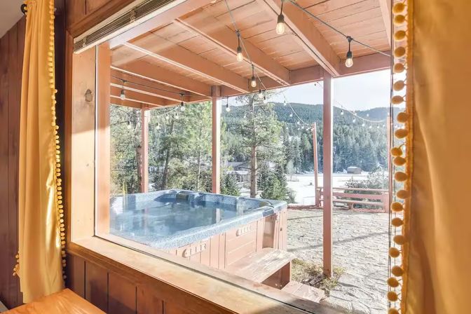 Cozy mountain cabin window framing an outdoor bubbling hot tub on a wooden deck with string lights, overlooking pine trees and a lakeside mountain view.