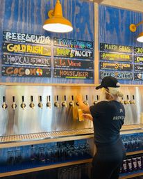 Person pouring a hazy beer from a stainless steel tap wall under yellow pendant lights in a craft brewery taproom with colorful chalkboard beer menu and rows of glassware.