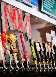 Close-up of colorful craft beer tap handles lined up on a bar draft system with brass faucets and a large red vintage letter sign in the background, ready to pour.