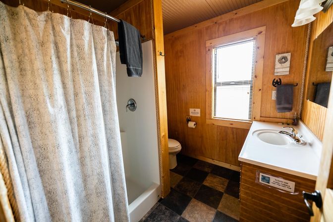 Cozy rustic cabin bathroom with wood-paneled walls, patterned shower curtain, single-sink vanity, toilet, window with blinds, and checkered tile floor.