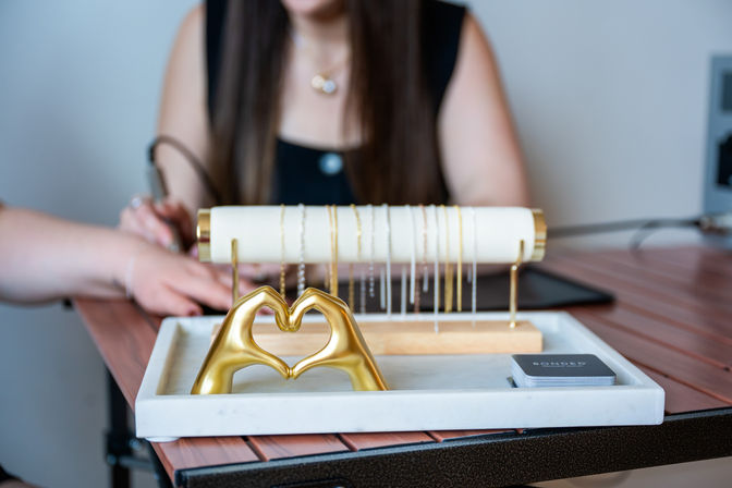 Close-up of a tabletop jewelry display with a marble tray holding a gold hands-formed heart sculpture and delicate gold and silver necklaces on a stand, blurred person working in the background in a boutique studio.