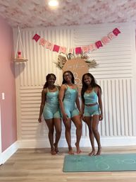 Three smiling women in matching teal activewear pose barefoot on a teal yoga mat in a pastel pink-and-white indoor studio decorated with a "Happy Birthday" banner and round floral sign.