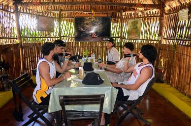 Group of friends drinking bottled beer at a green-checkered table inside a thatched-roof bamboo palapa, casual summer clothes and tropical outdoor dining vibe