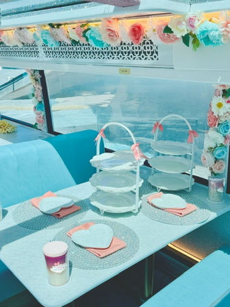 Pastel turquoise booth set for a whimsical seaside tea party with floral garlands, two white three-tier cake stands, heart-shaped plates on pink napkins, glitter placemats and a waterfront view.