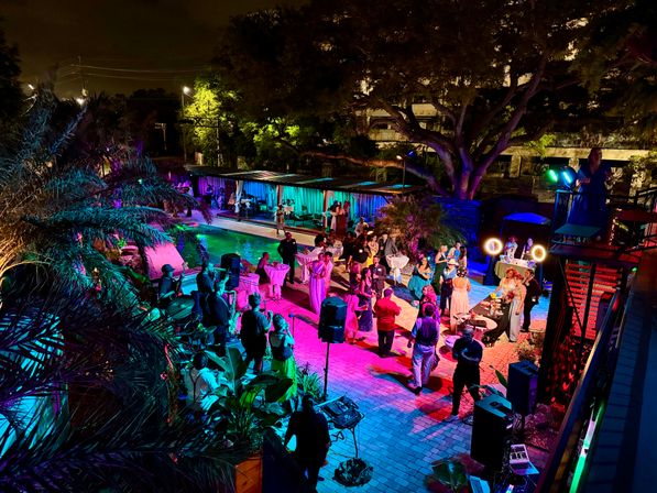 Vibrant poolside night party with colorful LED lighting, live band, and guests dancing and mingling under large tropical trees