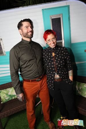 Two smiling adults posing in front of a turquoise retro camper backdrop at an outdoor event — man in olive shirt and rust pants, person with short red hair, dotted face makeup, floral black shirt and bolo tie.