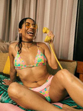 Laughing woman in a colorful floral bikini and yellow sunglasses holding a retro yellow telephone handset while lounging on a tropical-print bed in a bright resort-style room