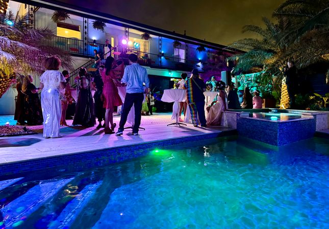 Vibrant poolside evening party in a tropical courtyard — guests in cocktail and formal attire mingling and dancing under colorful LED lights beside a lit balcony and palm trees.