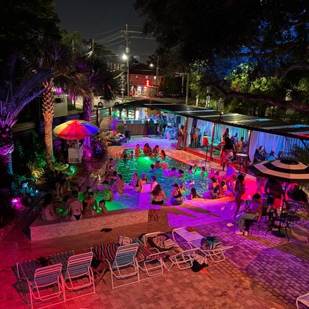 Vibrant nighttime outdoor pool party with multicolored LED lights, palm trees, cabanas and lounge chairs, groups socializing in and around an illuminated urban pool