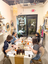 Cozy boutique craft studio candle-making workshop with four participants at a decorated table, rows of scent bottles, jars and pumpkins, macramé hanging décor and storefront door in the background