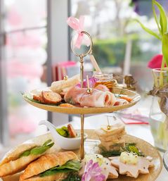 Delightful afternoon tea on a two-tier gold stand with croissant sandwiches, prosciutto, finger sandwiches, honey jar and floral garnish on a sunlit café table