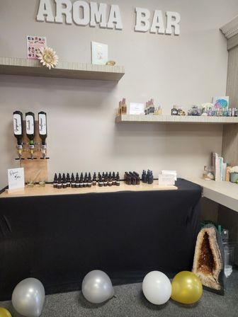 Aroma bar display in a wellness studio: wall sign and shelves with essential oils, three dispenser bottles, rows of amber dropper and spray bottles on a black-draped counter, small product displays, an amethyst geode and silver and gold balloons on the floor — aromatherapy retail setup.