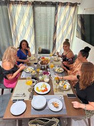 Five women enjoying a bright home brunch around a wooden dining table with eggs, fresh fruit, pastries, coffee and mimosas, patterned curtains and wall-mounted TV in the background.
