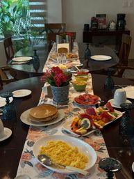 Indoor dining table set for breakfast with pancakes, scrambled eggs, colorful fruit platter, sliced tomatoes, cereal bowls and pastries, a coffee station in the background, floral centerpiece and patterned table runner.
