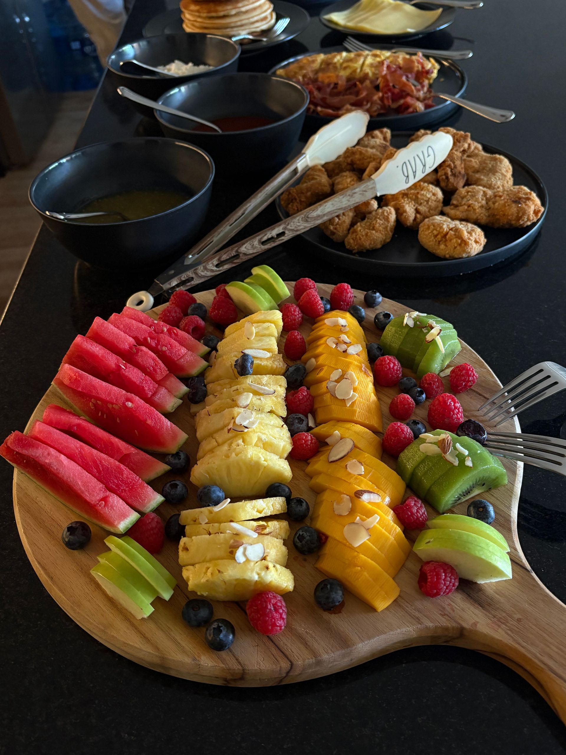 Vibrant brunch fruit platter on a wooden board — sliced watermelon, pineapple, mango, kiwi and green apple with raspberries, blueberries and almond flakes, breakfast buffet with fried chicken and pancakes in background