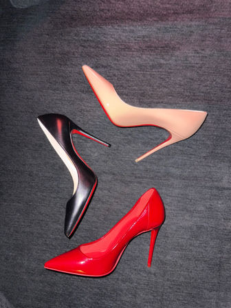 Three women's stiletto pumps—red patent, black, and nude—arranged on gray fabric background, glossy pointed-toe high heels with thin stiletto heels.