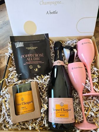 Luxury rosé champagne gift box with a bottle of rosé, boxed brut, two pink champagne flutes, gourmet rosé popcorn and chocolates nestled in gold-and-white shredded paper.