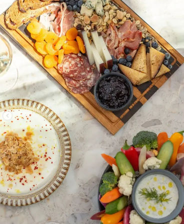 Top-down view of a colorful charcuterie and crudités spread on a marble tabletop — wooden board with cheeses, salami, prosciutto, dried apricots, nuts, blueberries and jam; bowl of fresh veggies with creamy dip and a yogurt plate topped with toasted almonds.