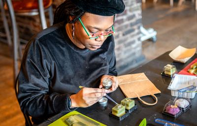 Artist in a beret and colorful glasses hand-decorating small square green handmade soaps or confections with tweezers at an indoor craft studio table with packaging and tools.